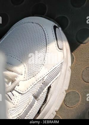 Closeup of a white sneaker with textured mesh and laces, resting on a rubber surface with circular pattern, illuminated by sunlight with soft shadows Stock Photo