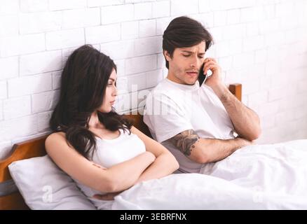 Couple Sitting in Bed With One Partner on the Phone While the Other Appears Upset Stock Photo