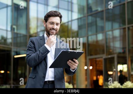 Businessman smiling confidently while holding a clipboard and reviewing important documents, reflecting successful planning and strategic thinking in Stock Photo