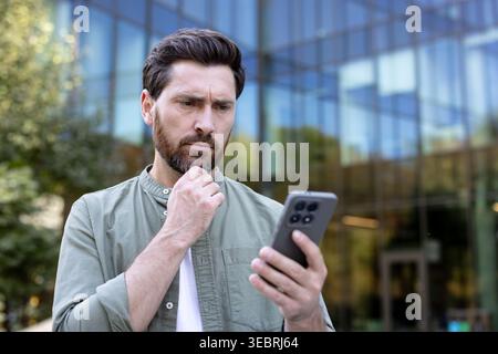 Serious man standing outdoors, contemplating news or a message on his smartphone, reflecting on important information in front of a modern office buil Stock Photo