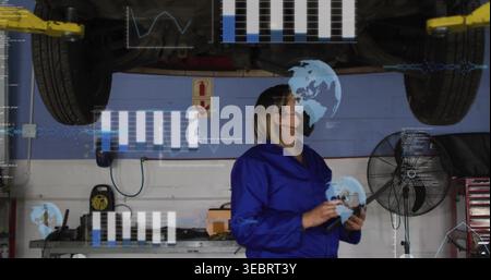 Inspecting tech blue coveralls black mask holding tablet under lifted vehicle in shop with overlays Stock Photo