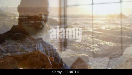 Framing, man wearing glasses and blue striped shirt holding camera at seaside window panes, sunset Stock Photo