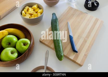 Long green cucumber lying on wooden cutting board on white counter, showing blue-handled knife Stock Photo