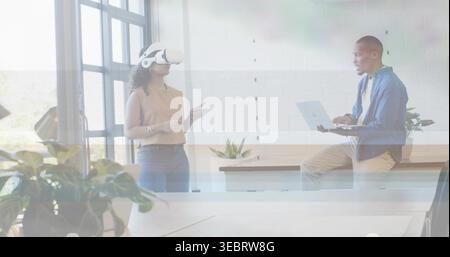 Demonstrating woman wearing top and white VR headset, man holding laptop at office, potted plants Stock Photo