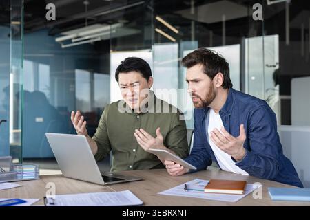 Two diverse businessmen feeling angry and confused while collaborating and facing a severe technical error or problem, on a laptop with documents on t Stock Photo