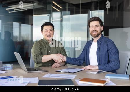 Two smiling multi-ethnic businessmen are shaking hands across a conference table in a modern office, signifying a successful partnership and agreement Stock Photo
