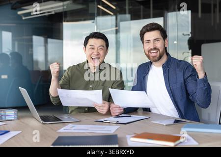 Diverse business partners feeling excited and raising fists in triumph, celebrating a successful project completion or achievement while holding docum Stock Photo