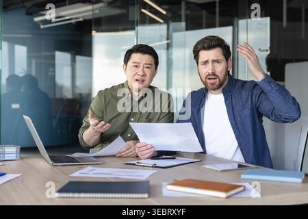 Two diverse businessmen sitting at a modern office table, expressing confusion and frustration while reviewing documents and discussing challenges dur Stock Photo