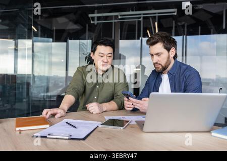 Two diverse business professionals are collaborating in a modern office, one showing content on his smartphone to the other, actively discussing start Stock Photo