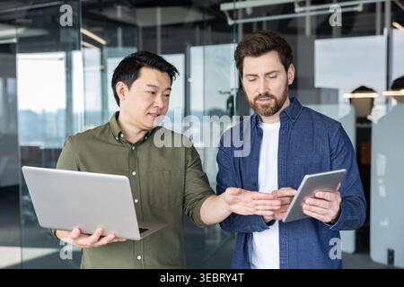 Two diverse business partners collaborating on a new work project, exchanging ideas and analyzing data using a laptop and digital tablet in a contempo Stock Photo