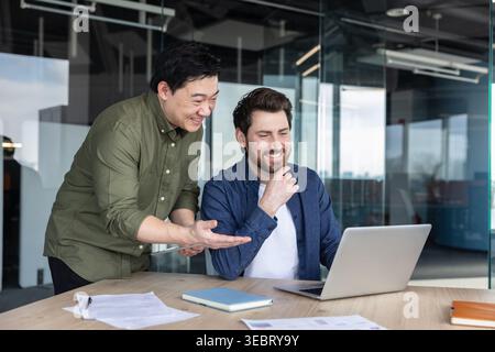 Two smiling multiethnic businessmen collaborate at a coworking desk, discussing data and sharing ideas while reviewing charts on a laptop in a modern Stock Photo