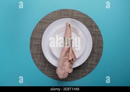 Closeup shows place setting on blue background. Soft, diffused lighting highlights pale pink damask napkin, silver napkin ring, white dishes, textured Stock Photo