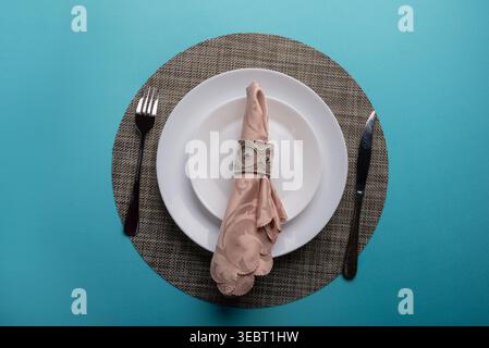 Overhead shot features pastel colored napkin, ornamental ring, white plates, metal fork, knife. Soft, diffused light highlights textures. Represents d Stock Photo
