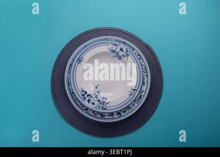 Overhead shot on turquoise backdrop presents plates stacked on matching woven placemat. Plates feature blue floral pattern. Soft, diffused lighting en Stock Photo