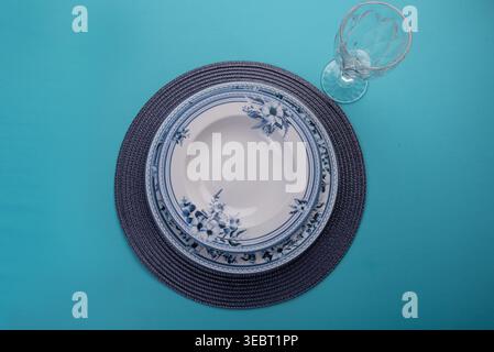 Overhead shot featuring stacked porcelain plates, floral design, sits atop woven placemat alongside crystal water glass. Soft studio light, textures e Stock Photo