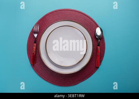 Overhead shot featuring empty white plates set on red woven placemat, minimalist setup. Background in light blue. Silver cutlery on the side. Studio l Stock Photo