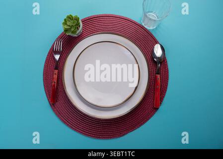 Overhead shot of minimalist place setting on turquoise table. Includes white plates, silver cutlery, succulent, and glass. Soft, even lighting. Ideal Stock Photo