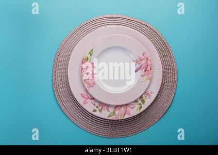 Overhead shot features pink floral patterned plates on woven placemat, atop of vibrant clear blue background. Soft indirect lighting. Ideal for lifest Stock Photo