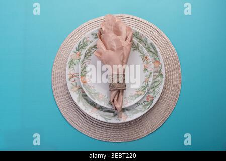 Overhead shot displays floral plates, neatly folded napkin, decorative ring on textured woven placemat above azure table. Soft studio lighting, neutra Stock Photo