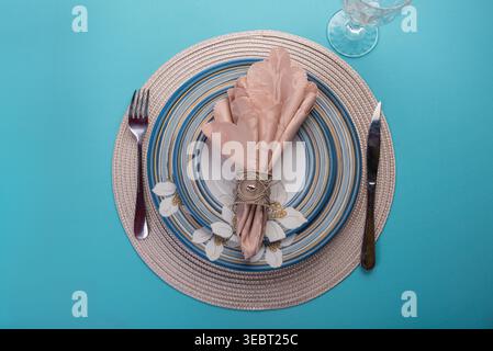 Overhead shot of table setting. Features floral china, pink napkin, ornate ring, silverware on round woven placemat. Bright, even lighting, blue backg Stock Photo