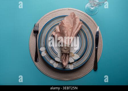 Overhead shot of formal dinning setup. Features blue striped dinnerware, pink napkin, decorative silver ring, modern flatware on woven charger, set on Stock Photo