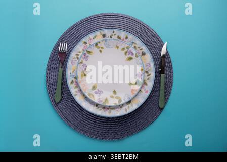 Still life of floral patterned plates on a woven mat. Light enhances textures. Suggests healthy dining, wellness and simple beauty. Blue background ad Stock Photo