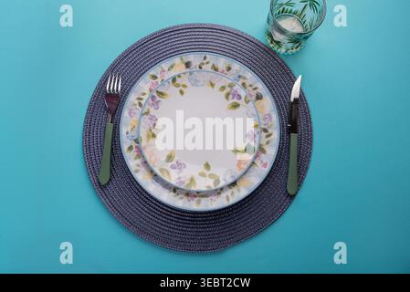 Overhead arrangement features floral patterned plates, glass cup and cutlery, isolated on vibrant turquoise backdrop. Studio shot with soft light, ide Stock Photo