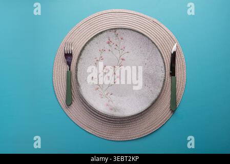 Overhead shot of plate with floral pattern, on placemat with knife and fork. Table setting features soft, diffused lighting and a textured background. Stock Photo