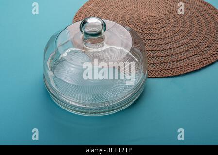 Glass cloche in a studio shot, placed on a vibrant turquoise surface, beside a rustic brown woven placemat. Soft lighting highlights glass textures. M Stock Photo