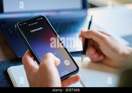 Hand holds smartphone with unknown incoming call above open notebook at home desk. Concept of work interruption, spam call and unproductivity at works Stock Photo