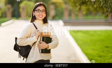 Student Enjoys a Sunny Day Outdoors While Holding Academic Materials and Wearing Wireless Earbuds Stock Photo