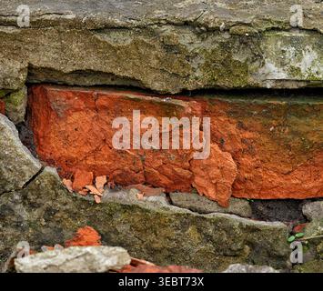 A close-up shot of a weathered and crumbling red brick embedded in a cracked concrete structure with traces of green moss. Close up. Stock Photo