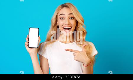 Young Woman Shows Phone and Smiles in a Bright Setting With a Blue Background Stock Photo