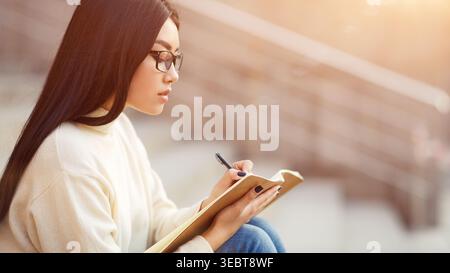 Young Woman Writing in Notebook While Sitting Outdoors in Soft Sunlight Stock Photo