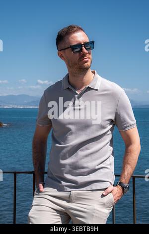 Man in light gray polo by railing with sea and Antibes coastline Stock Photo