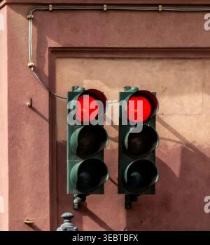 Two red traffic lights mounted on a textured terracotta building wall. Electrical cables and sharp shadows are visible in bright daylight. Stock Photo