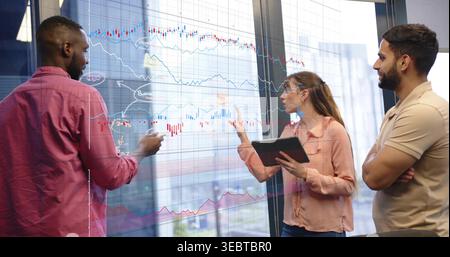 Discussing three pros pointing at glass wall charts in office with tablet, wearing red pink beige Stock Photo