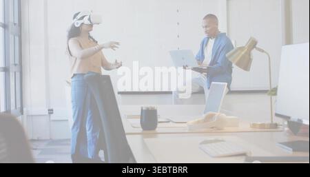 Interacting woman with white VR headset, sleeveless top, gesturing in open office, laptop on desk Stock Photo