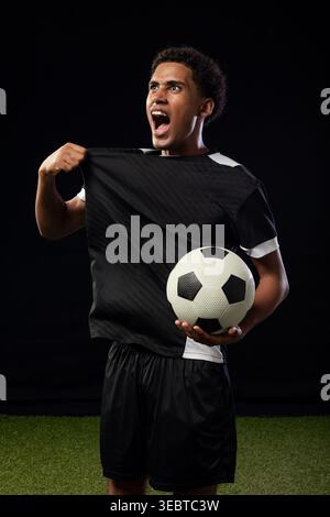African American man in black soccer kit shouting and holding black-and-white ball on studio turf Stock Photo