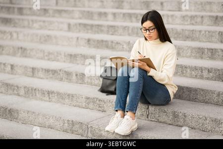 Young Woman Studying on Stone Steps While Taking Notes During a Sunny Afternoon Stock Photo