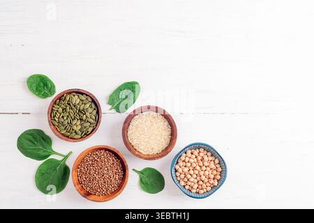 Cereals and seeds in bowls and green spinach leaves on white wooden table. Rice, buckwheat, chickpeas and pumpkin seeds. Top view, flat lay, copy spac Stock Photo