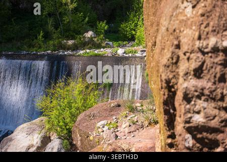 Even sheet of water spills over a low concrete weir in Madeira, with sunlit boulders, moss, and subtropical plants. Midday light highlights rock textu Stock Photo