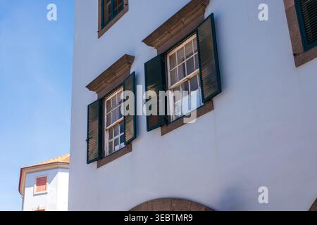 Whitewashed facade with green wooden shutters and paned windows in Funchal, Madeira. Strong sunlight casts sharp shadows on textured masonry and an ar Stock Photo