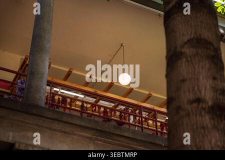 Low angle view shows a balcony railing, a round pendant light on a beige ceiling, wooden beams, concrete pillars, and a tree trunk in a semi open stru Stock Photo