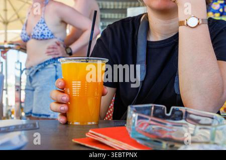 Young woman sipping orange juice through straw at beach cafe with bikini beachgoers in background. Paphos, Cyprus by the Mediterranean sea, Stock Photo