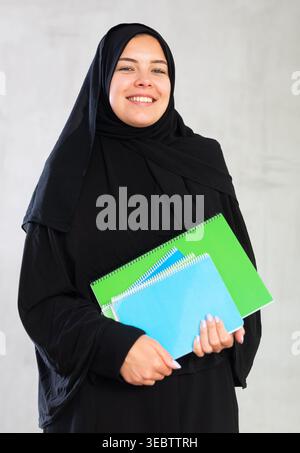 Smiling young female student dressed in traditional islamic clothes holding textbooks Stock Photo