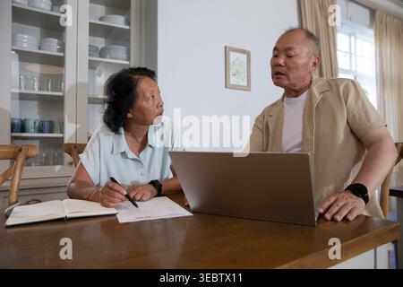 Asian couple with senior woman and mature man discussing paperwork at kitchen table using laptop Stock Photo