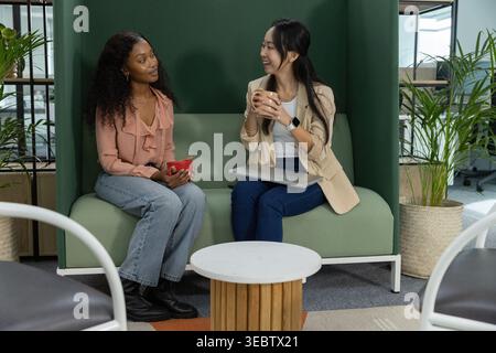 Diverse female coworkers sitting on green booth sofa in office, holding red mug and closed laptop Stock Photo