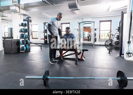 African American male trainer and client reviewing tablet while sitting on red bench in gym. Barbell, weights, plates, mirrors, studio, fitness, indus Stock Photo