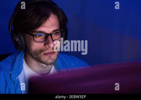 Young Man Focused on Computer Screen in Low Light Environment Wearing Headphones Stock Photo